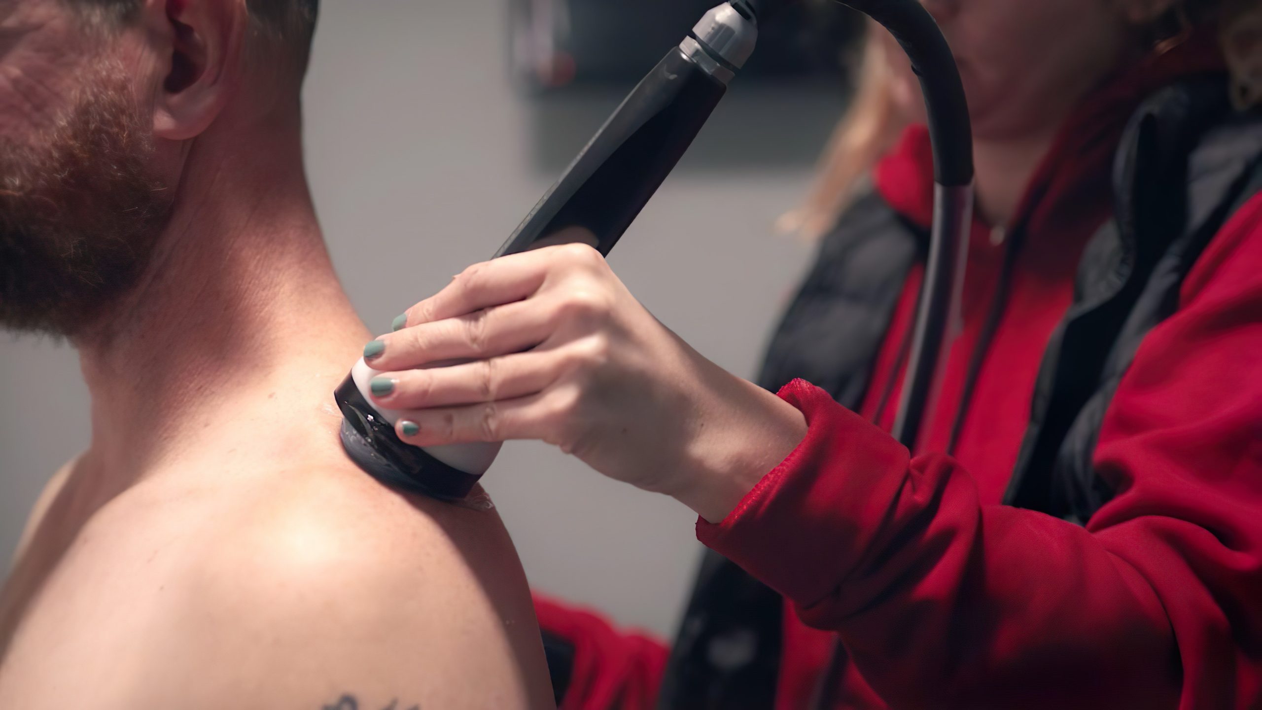 Healthcare worker in a red jacket uses a handheld device on a shirtless man's neck during a medical examination/assessment.