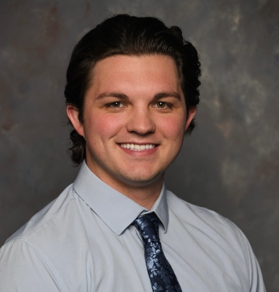 Smiling man in a light blue dress shirt and patterned navy tie against a neutral studio backdrop.