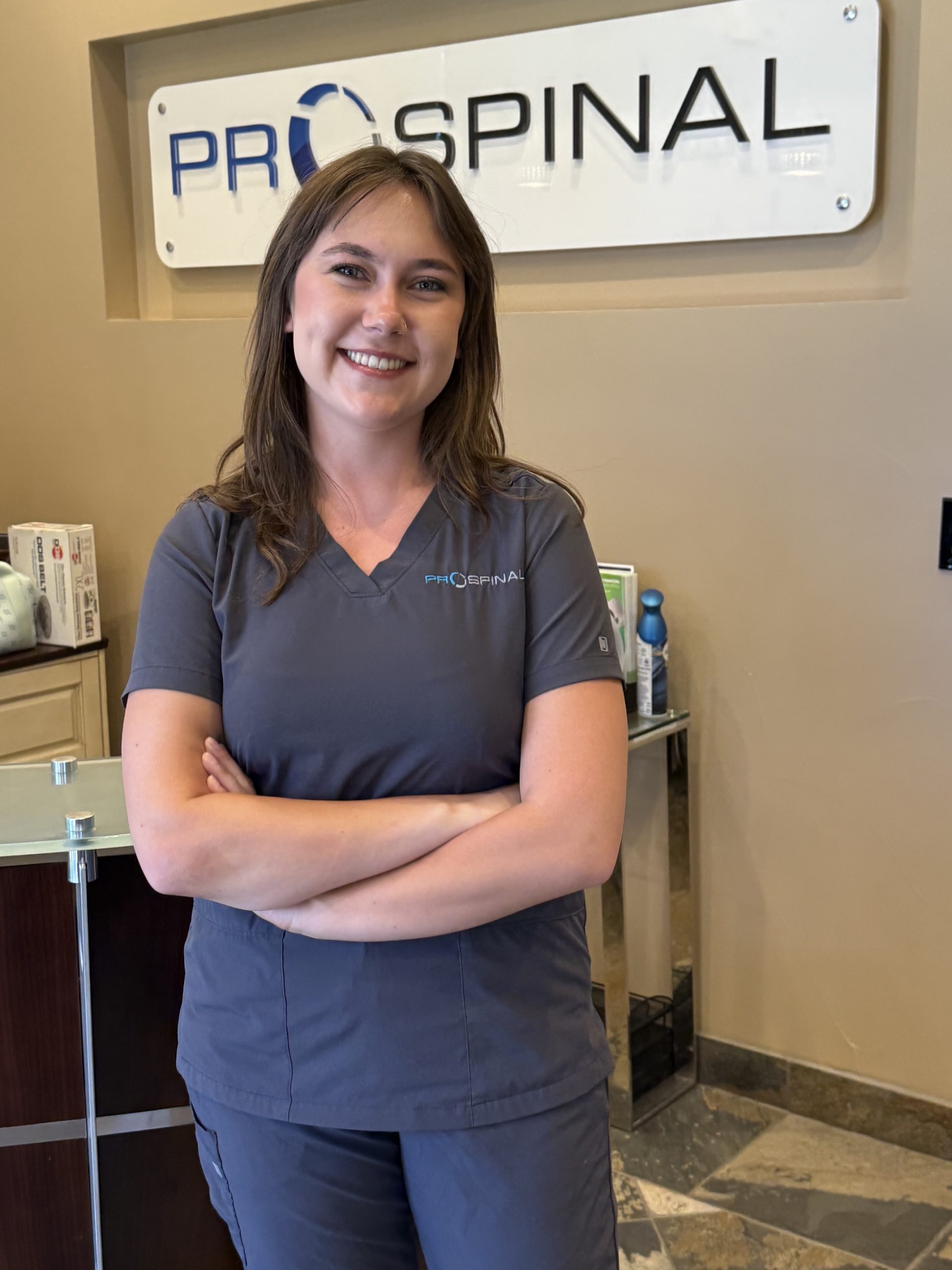 Smiling female healthcare worker in gray scrubs standing in front of a reception sign that reads PROSPINAL.